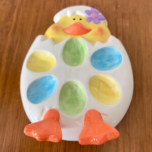 K&ouml;nnte beinhalten: Ein Keramik-Ostereierhalter in Form eines K&uuml;kens, das aus einem Ei schl&uuml;pft. Das K&uuml;ken ist gelb mit einem orangen Schnabel und einer lila Blume. Das Ei hat Aussparungen f&uuml;r bunte Eier.