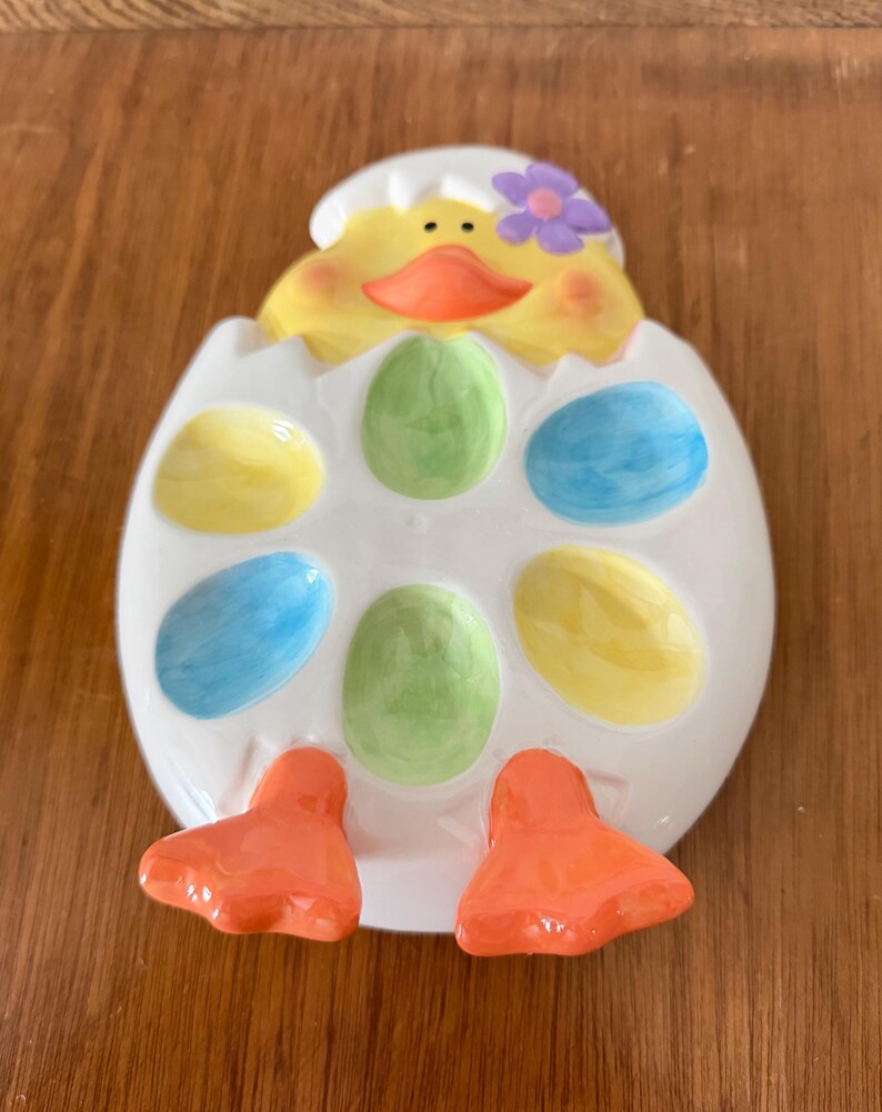 K&ouml;nnte beinhalten: Ein Keramik-Ostereierhalter in Form eines K&uuml;kens, das aus einem Ei schl&uuml;pft. Das K&uuml;ken ist gelb mit einem orangen Schnabel und einer lila Blume. Das Ei hat Aussparungen f&uuml;r bunte Eier.