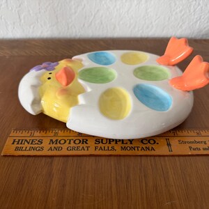K&ouml;nnte beinhalten: Eine keramische, eif&ouml;rmige Osterschale mit einem gelben K&uuml;ken, das aus einer zerbrochenen Eierschale kommt. Die Schale hat sechs farbige, eif&ouml;rmige Vertiefungen. Orangefarbene F&uuml;&szlig;e ragen von der Seite heraus. Die Schale steht auf einer Holzoberfl&auml;che.