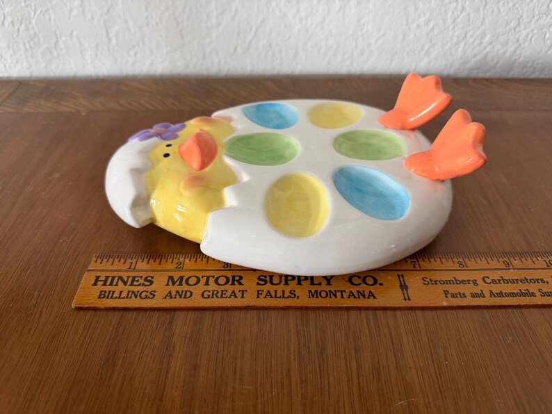 K&ouml;nnte beinhalten: Eine keramische, eif&ouml;rmige Osterschale mit einem gelben K&uuml;ken, das aus einer zerbrochenen Eierschale kommt. Die Schale hat sechs farbige, eif&ouml;rmige Vertiefungen. Orangefarbene F&uuml;&szlig;e ragen von der Seite heraus. Die Schale steht auf einer Holzoberfl&auml;che.