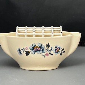 May include: A white ceramic flower vase with a floral design in blue, pink, and green. The vase has a built-in metal grid for holding flowers.