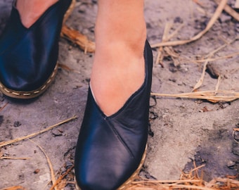 May include: A pair of black leather slip-on shoes with a pointed toe and a v-shaped opening at the top. The shoes are worn by a person standing on a dirt path with dry grass.