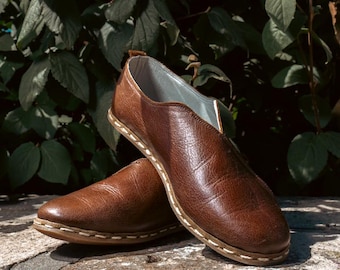 May include: A pair of brown leather slip-on shoes with white stitching around the edges. The shoes have a pointed toe and a flat sole.