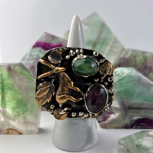 Peut inclure: Bague carrée avec une base en métal foncé, ornée de motifs floraux et animaliers dorés. Elle comporte une pierre verte et une pierre rouge. La bague est présentée sur un support blanc, avec des cristaux colorés en arrière-plan.