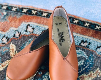 May include: A pair of brown leather slip-on shoes with a pointed toe and a v-shaped opening. The shoes are on a patterned rug and the size 39 W is visible on the inside of one shoe.