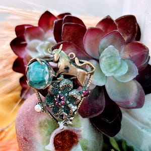 May include: A detailed shot of a brooch featuring a gold bull, a teal stone, and a floral design with purple accents. The brooch is set against a backdrop of burgundy and green succulents and a peach-colored fruit.