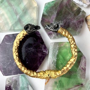 May include: A gold-colored bracelet with textured detailing and ram's head accents. The bracelet is set against a backdrop of colorful, geometric-shaped stones in shades of purple, green, and white.