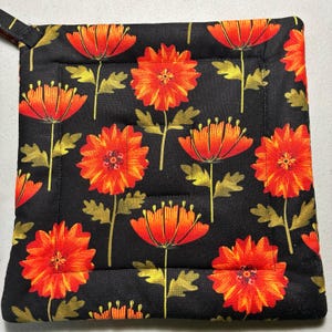 May include: A square quilted pot holder featuring a black background with a repeating pattern of vibrant orange and red flowers with olive green leaves and stems. The fabric is stitched with a square border pattern and includes a small corner loop.