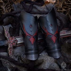 May include: Two black leather arm guards with red flame designs and diamond details. The guards are laced with black cord and are displayed on a fur-like surface. A decorative sword is also visible.