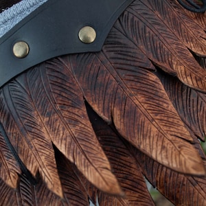 May include: A brown leather belt with a decorative feather design. The feathers are carved into the leather and have a dark brown color. The belt has a black leather strap with a metal buckle.