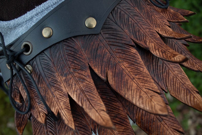 May include: A brown leather belt with a decorative feather design. The feathers are carved into the leather and have a dark brown color. The belt has a black leather strap with a metal buckle.