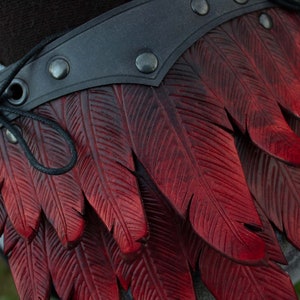 May include: A close-up of a red leather belt with a feather design. The belt is made of dark brown leather and has a black strap with metal rivets. The feathers are carved into the leather and are a deep red color.