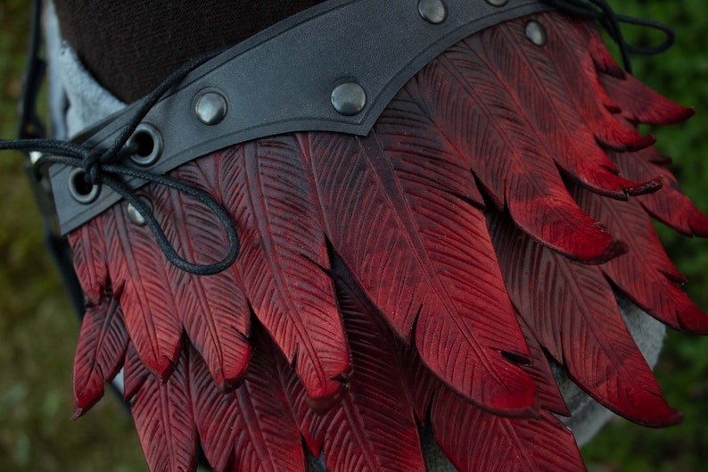 May include: A close-up of a red leather belt with a feather design. The belt is made of dark brown leather and has a black strap with metal rivets. The feathers are carved into the leather and are a deep red color.