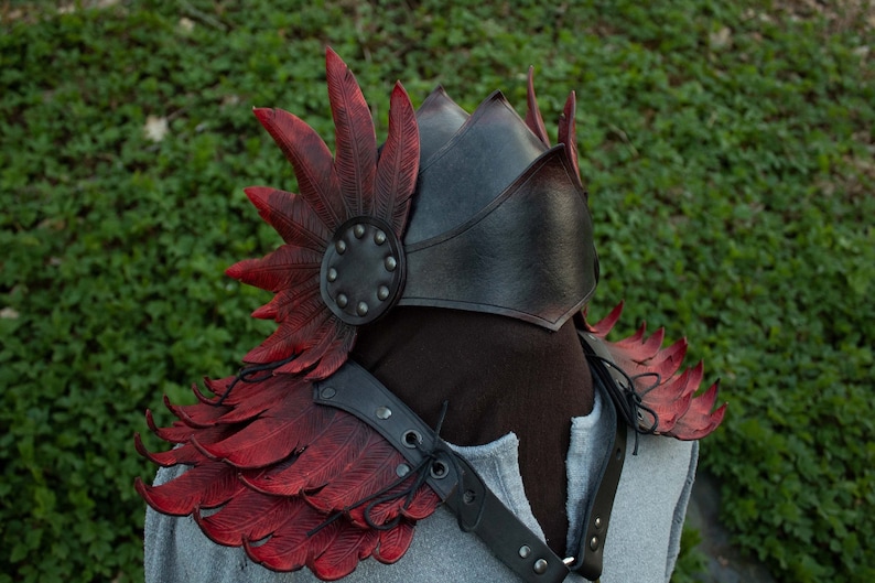 May include: A black leather shoulder armor with red leather feathers. The armor has a decorative design with rivets and straps.