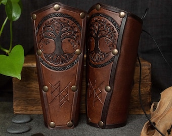 May include: Two brown leather arm guards with a tree of life design. The arm guards have brass rivets and a dark brown border. The tree of life is embossed in the center of each guard. The bottom of each guard has a geometric design.