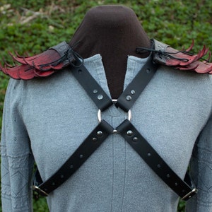 May include: Red leather shoulder armor with black straps and a silver ring. The armor is designed to look like wings with spikes.