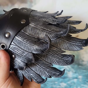 May include: A black leather cuff with silver-colored feathers. The cuff has a row of silver-colored studs along the edge.