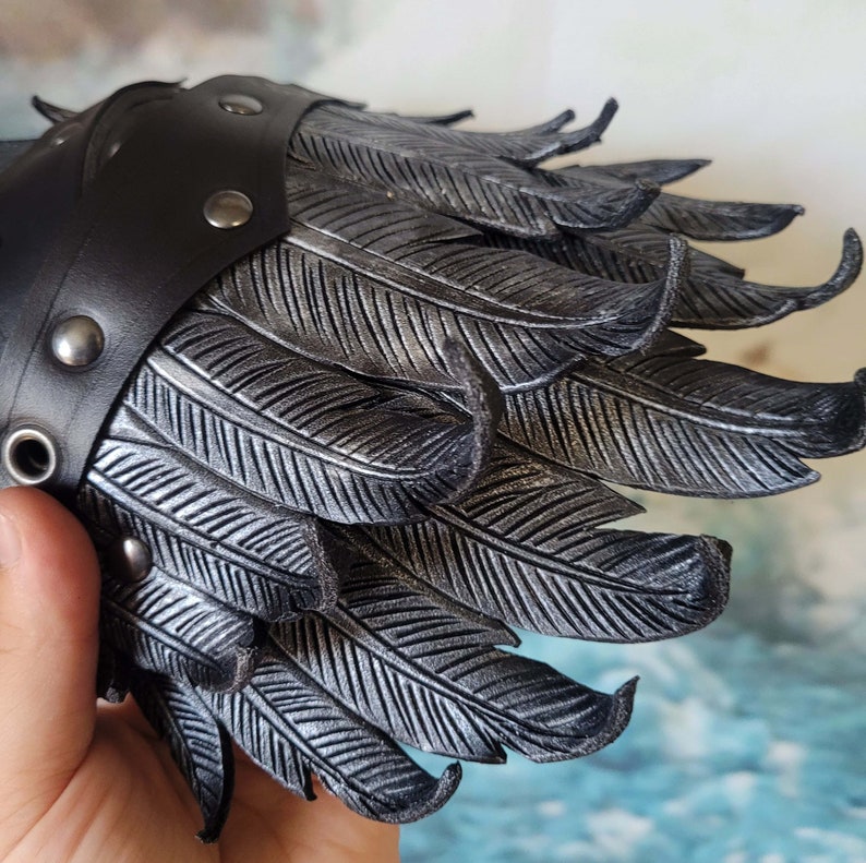 May include: A black leather cuff with silver-colored feathers. The cuff has a row of silver-colored studs along the edge.