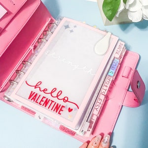 May include: Pink leather bound planner with a clear plastic cover and the words "hello valentine" in red. The planner has tabs with the words "to order", "lilta", "etsy", "results", "goals", and "inbox".