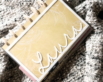 May include: A beige and white planner with a floral design and the name "Laura" written in white cursive on the front. The planner has a clear plastic cover and a white ring binder.
