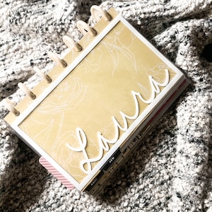 May include: A beige and white planner with a floral design and the name "Laura" written in white cursive on the front. The planner has a clear plastic cover and a white ring binder.