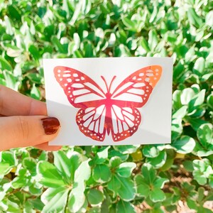 May include: A red iridescent butterfly sticker on a white background. The butterfly has a detailed design with intricate wing patterns.