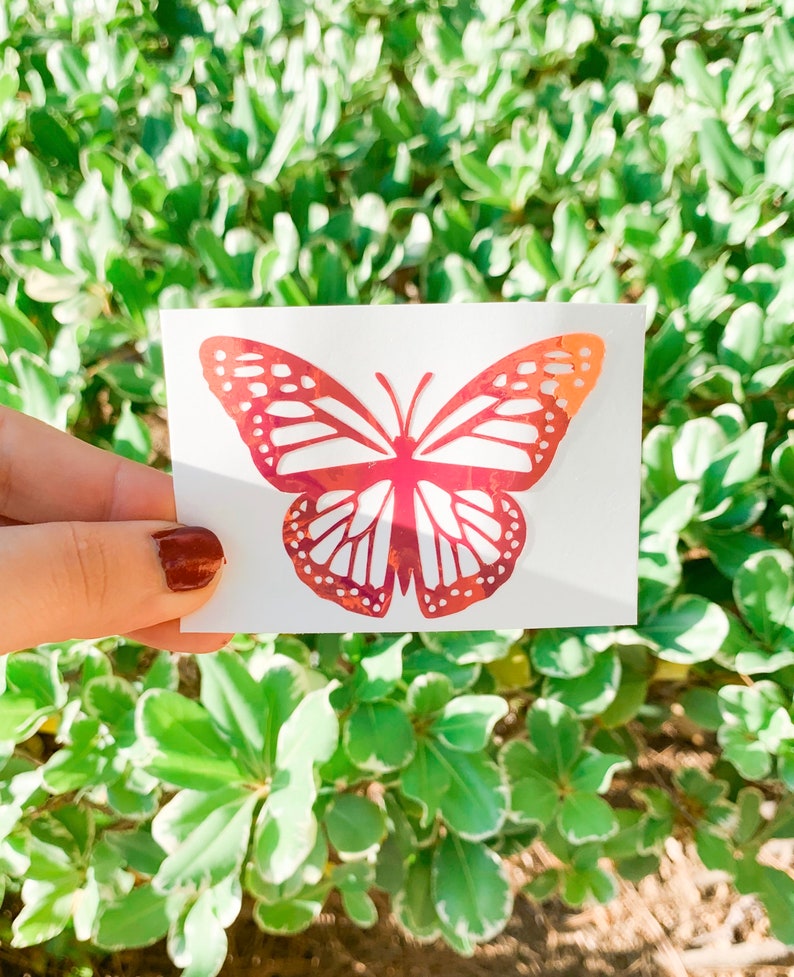 May include: A red iridescent butterfly sticker on a white background. The butterfly has a detailed design with intricate wing patterns.