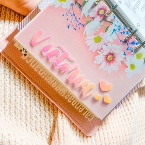 Può includere: Un planner rosa con motivo floreale e una copertina in plastica trasparente. La copertina ha un adesivo in vinile rosa iridescente che dice "Victoria" con due piccoli cuori rosa.