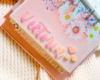 May include: A pink floral patterned planner with a clear plastic cover. The cover has a pink iridescent vinyl sticker that says "Victoria" with two small pink hearts.