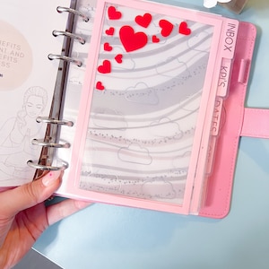 May include: A pink planner with a clear plastic pocket on the front. The pocket has a gray and white patterned background with red heart stickers. The planner has tabs labeled "Inbox", "KPI's", and "Dates".