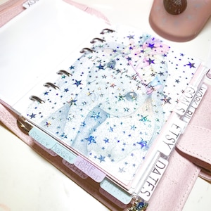 Pu&ograve; includere: Un'agenda rilegata in pelle rosa con una copertina in plastica trasparente con un motivo a stelle olografiche. L'agenda ha un sistema di rilegatura ad anelli argentati e include schede con le etichette "Date", "Etsy", "Fatture", "Cose da fare", "Date" e "Settimana a".