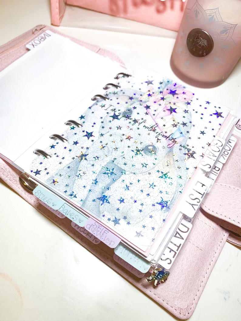 Pu&ograve; includere: Un'agenda rilegata in pelle rosa con una copertina in plastica trasparente con un motivo a stelle olografiche. L'agenda ha un sistema di rilegatura ad anelli argentati e include schede con le etichette "Date", "Etsy", "Fatture", "Cose da fare", "Date" e "Settimana a".