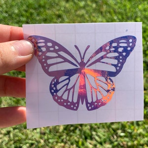 May include: A holographic butterfly sticker with iridescent purple and pink colors. The sticker is on a white sheet of paper with a grid pattern.