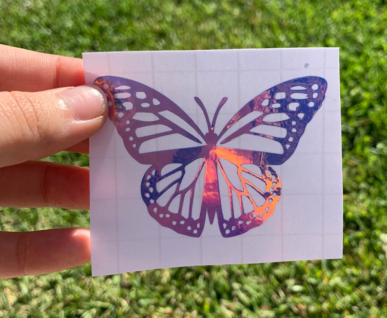 May include: A holographic butterfly sticker with iridescent purple and pink colors. The sticker is on a white sheet of paper with a grid pattern.