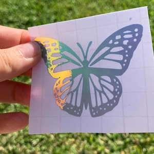 May include: A holographic butterfly sticker with a yellow, green, and blue iridescent finish. The sticker is on a white sheet with a grid pattern.