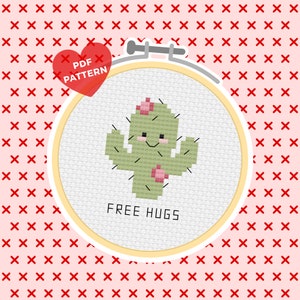 May include: A cross-stitch pattern featuring a green cactus with a pink flower on top and the text "FREE HUGS". The pattern is in a round embroidery hoop with a white background.