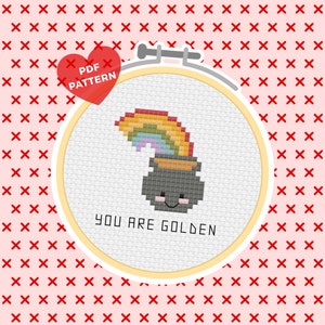 May include: A cross stitch pattern featuring a cartoon pot of gold with a rainbow on top. The pot of gold has a smiling face and the text "YOU ARE GOLDEN" is below it.