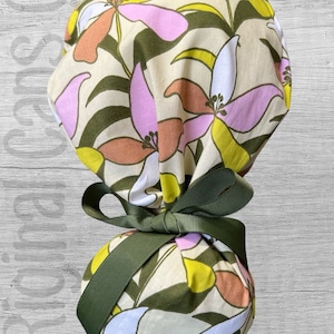 May include: A floral patterned scrub cap with a green ribbon. The cap features a cream background with large flowers in shades of pink, yellow, orange, and blue, along with green leaves. The ribbon is a solid olive green.