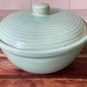 May include: A light blue ceramic bowl with a matching lid. The bowl and lid have a ridged design. The lid has a small, round handle. The bowl is sitting on a wooden surface.