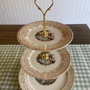 May include: A three-tiered serving stand with gold-colored metal handle and trim. The plates are cream-colored with a gold floral pattern and a central image of a couple in a garden setting. The stand is on a green and white checkered tablecloth.