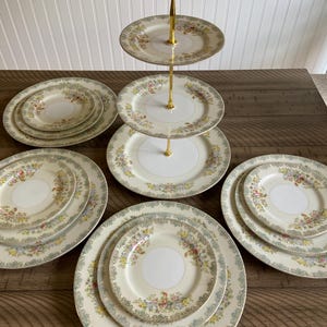 May include: A vintage porcelain tea set with floral patterns. The set includes a three-tiered serving stand with gold accents, and several plates of varying sizes. The plates feature a cream-colored background with a floral border.