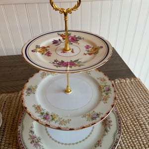 May include: A three-tiered serving stand with gold-colored hardware. Each tier is a decorative plate with floral patterns in shades of pink, yellow, and purple. The plates have a cream-colored background and are trimmed with gold and dark blue.