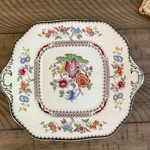 May include: Square, off-white serving plate with a green trim and handles. The plate features a central floral design in shades of pink, orange, blue, and yellow, surrounded by a brown and white border. Decorative tableware.