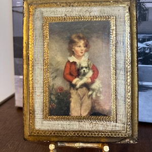 May include: A framed painting of a young boy in a red jacket holding a small white dog. The painting is on a gold easel.