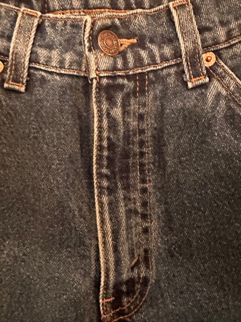 May include: Close-up of the front fly and waistband of a pair of dark blue denim jeans. The jeans have a button fly and belt loops.
