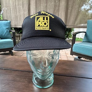 May include: Black trucker hat with a flat brim, featuring the text "War Wagon" and "ALL PRO AUTO PARTS" in gold lettering. The hat is displayed on a glass head. The hat has a mesh back.