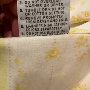 May include: A white care label with black text outlining washing instructions for a garment. The text reads: "MACHINE WASH - USE WARM SETTING. 2. DO NOT OVERLOAD WASHER OR DRYER. 3. TUMBLE DRY AT HOT OR COTTON SETTING. 4. REMOVE PROMPTLY FROM DRYER AND FOLD. 5. LAUNDER HIGH FASHION COLORS SEPARATELY. 6. DO NOT USE BLEACH ON DARK COLORS OR PRINTS."