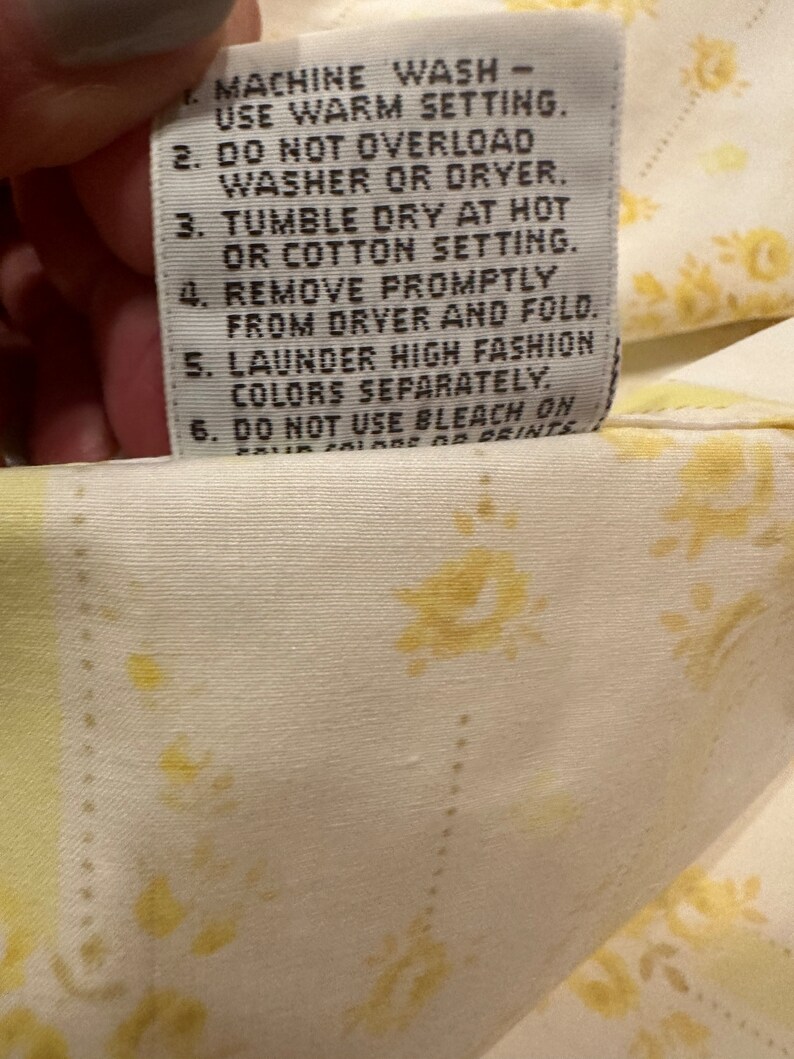 May include: A white care label with black text outlining washing instructions for a garment. The text reads: "MACHINE WASH - USE WARM SETTING. 2. DO NOT OVERLOAD WASHER OR DRYER. 3. TUMBLE DRY AT HOT OR COTTON SETTING. 4. REMOVE PROMPTLY FROM DRYER AND FOLD. 5. LAUNDER HIGH FASHION COLORS SEPARATELY. 6. DO NOT USE BLEACH ON DARK COLORS OR PRINTS."