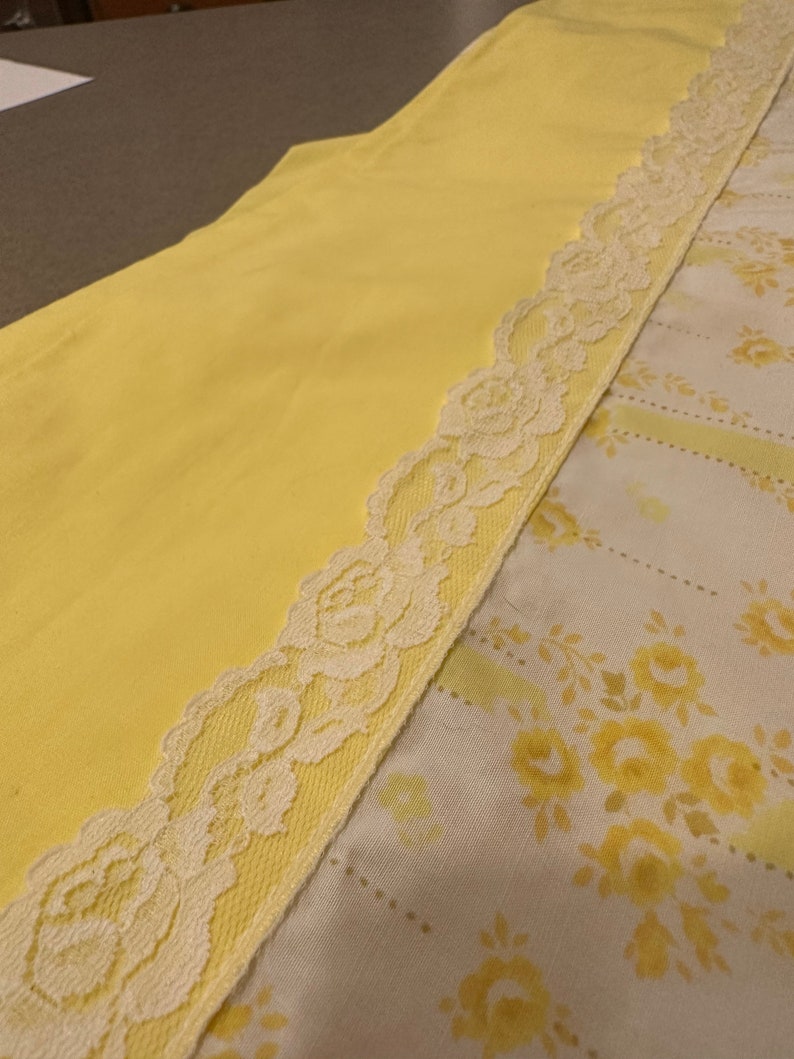 May include: A yellow fabric with a white lace trim. The lace has a floral pattern. The fabric is folded in half, with the lace trim on the outside.