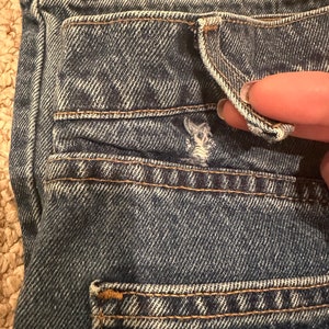 May include: Close-up of the back pocket of a pair of blue denim jeans. The pocket has a flap and is stitched with a contrasting thread.